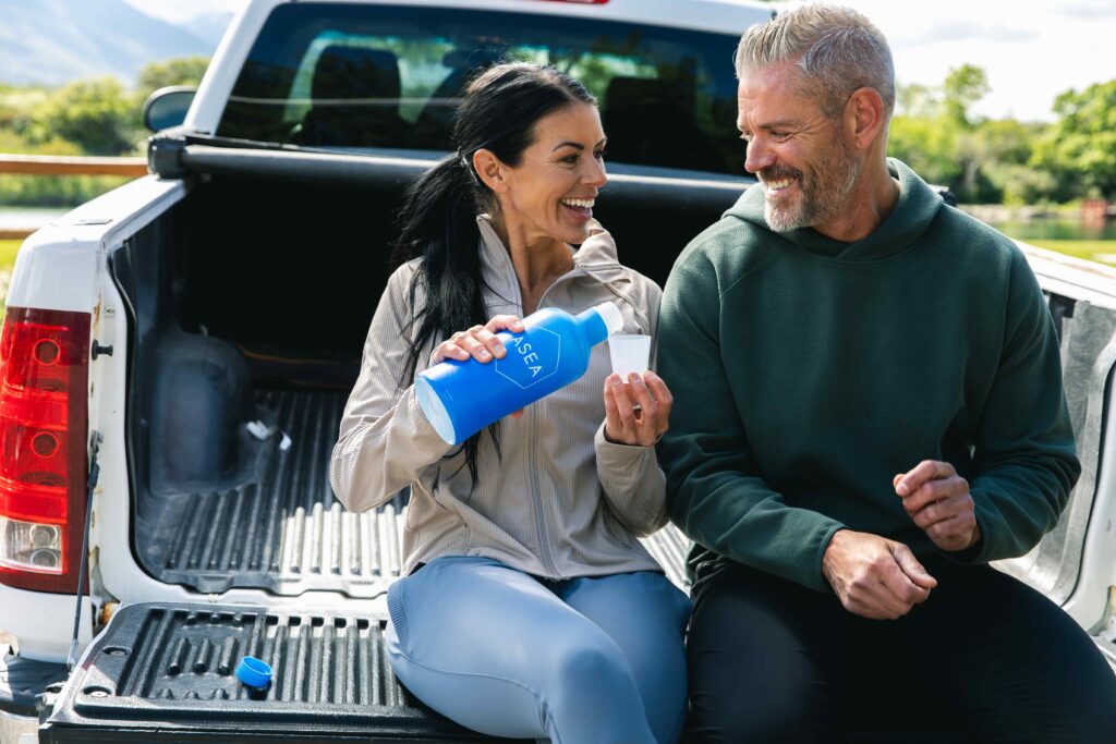 A man and woman sit on the back of a truck outdoors, smiling and holding an ASEA Redox Cell Signaling Supplement bottle as part of their daily wellness routine that supports cellular health and active living.