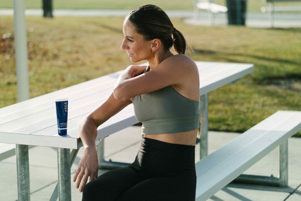 A woman stretches her shoulder after a workout while seated outdoors, with a muscle recovery gel nearby, representing post-workout muscle recovery support for women.