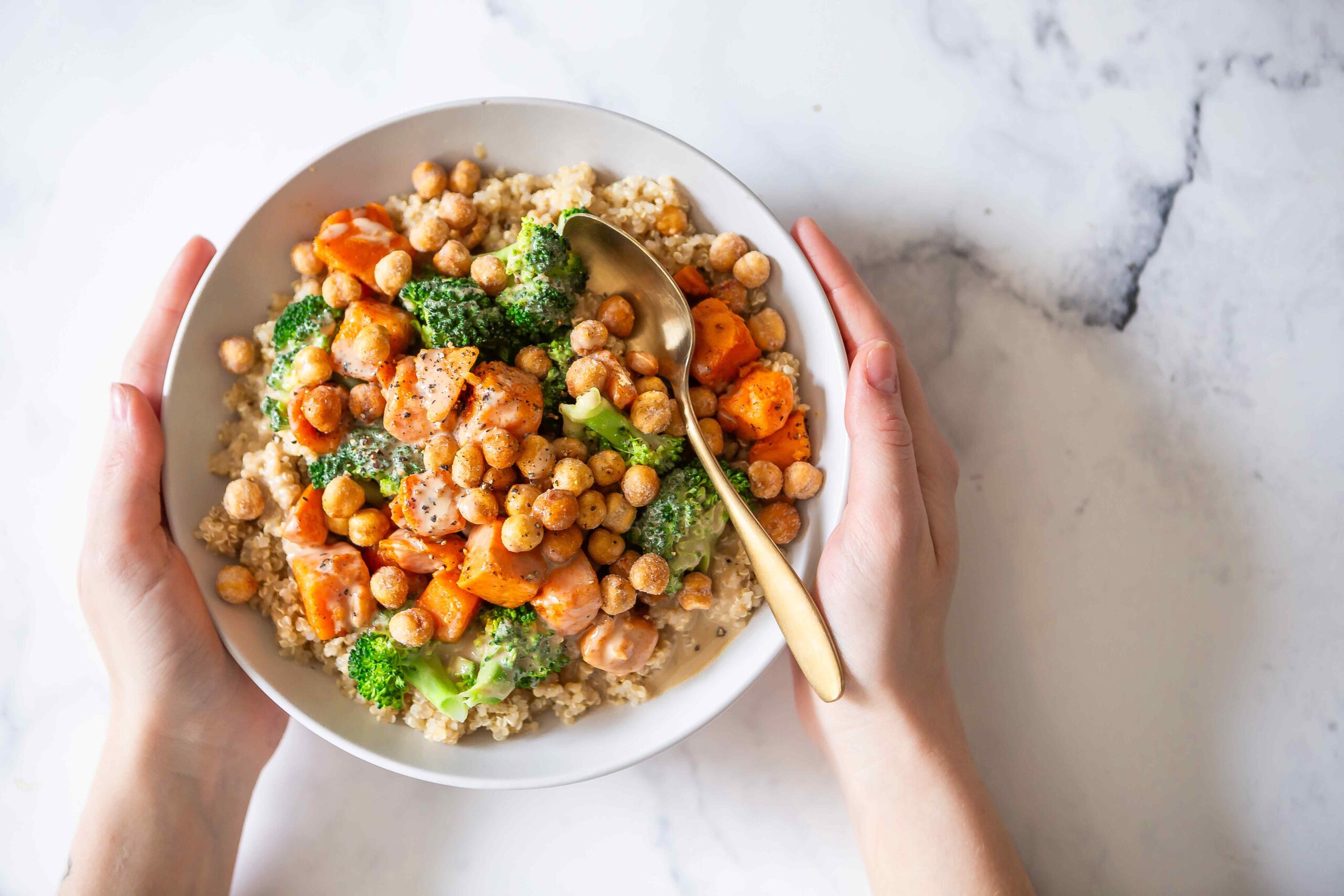 Hands hold a bowl filled with quinoa, roasted sweet potatoes, chickpeas, and broccoli on a bright countertop. It visually supports the post-workout recovery bowl recipe theme focused on nourishing whole foods.
