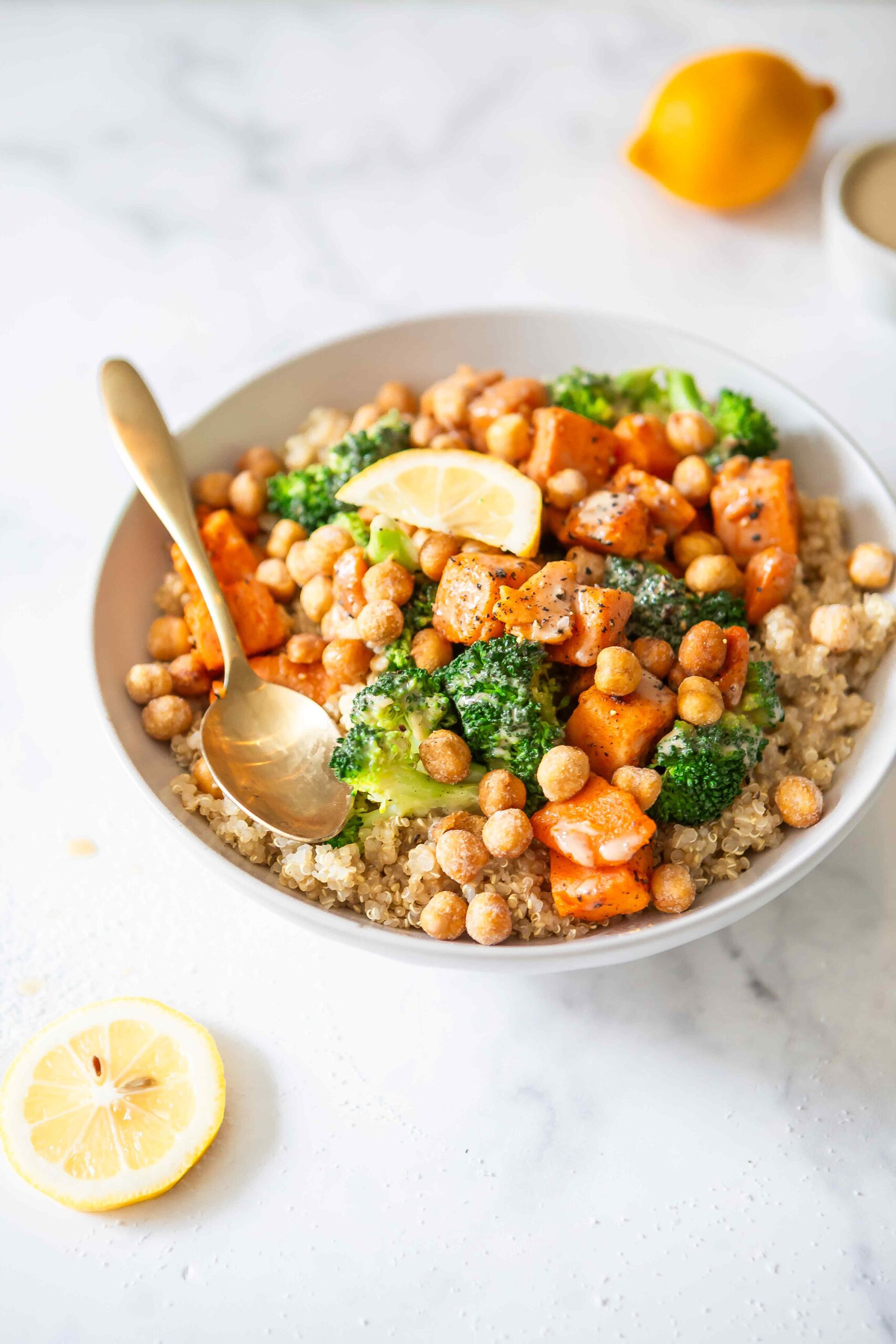 A plated quinoa bowl topped with chickpeas, broccoli, roasted sweet potatoes, and a lemon slice, styled with a spoon on a light background. This image represents the post-workout recovery bowl recipe featured in the article.
