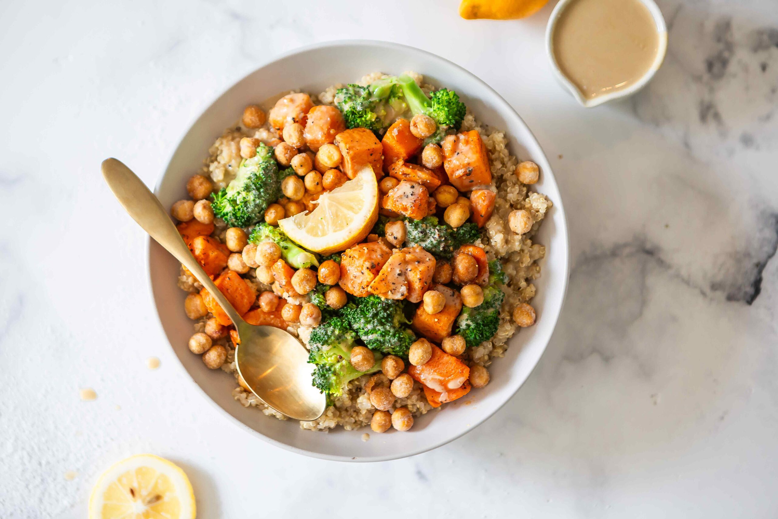 A close-up of a quinoa recovery bowl topped with roasted sweet potatoes, chickpeas, broccoli, and a lemon wedge on a light marble surface. This post-workout recovery bowl recipe highlights whole-food ingredients for balanced recovery nutrition.